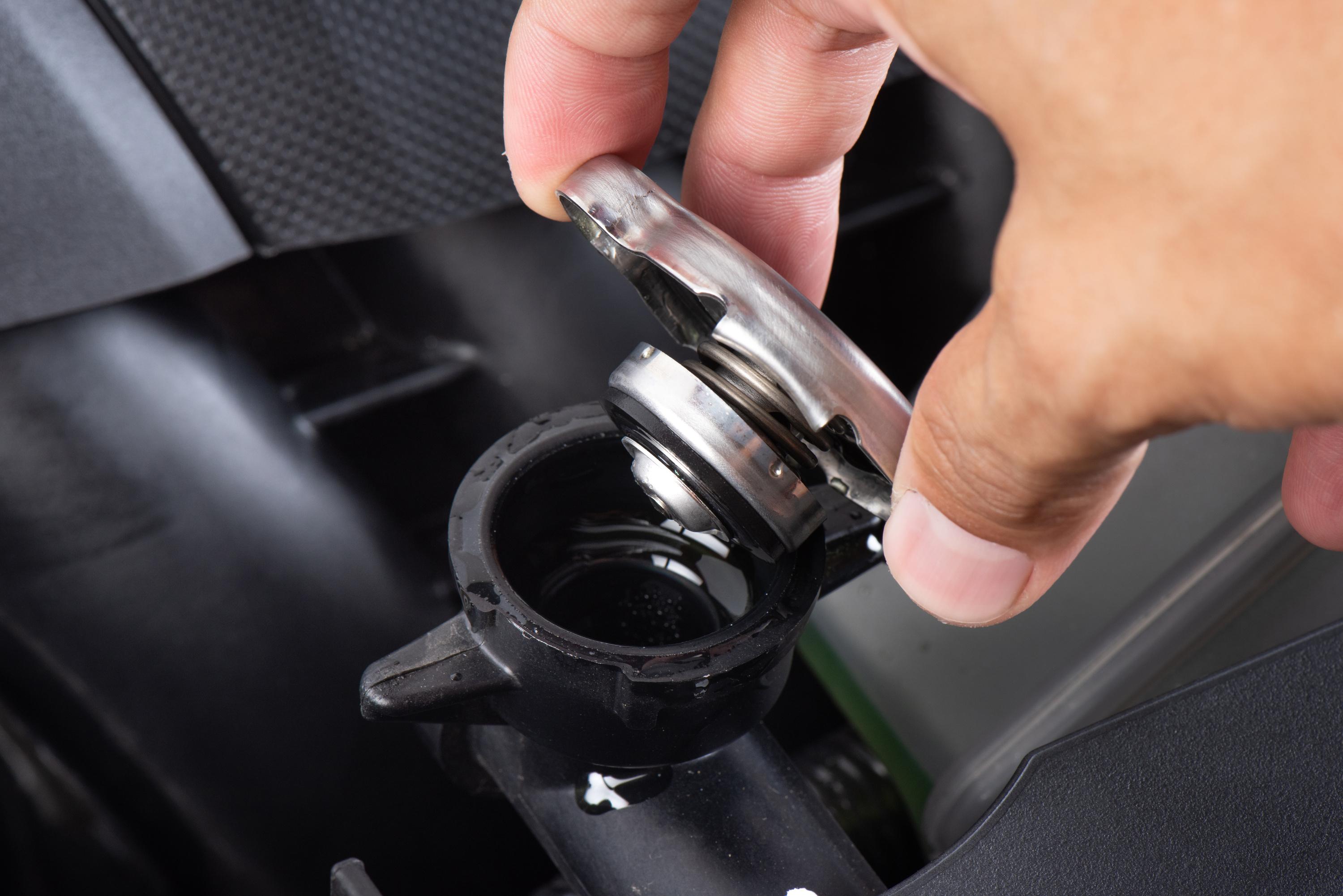 Hand removing a metal radiator cap with coolant visible inside the car's radiator opening.