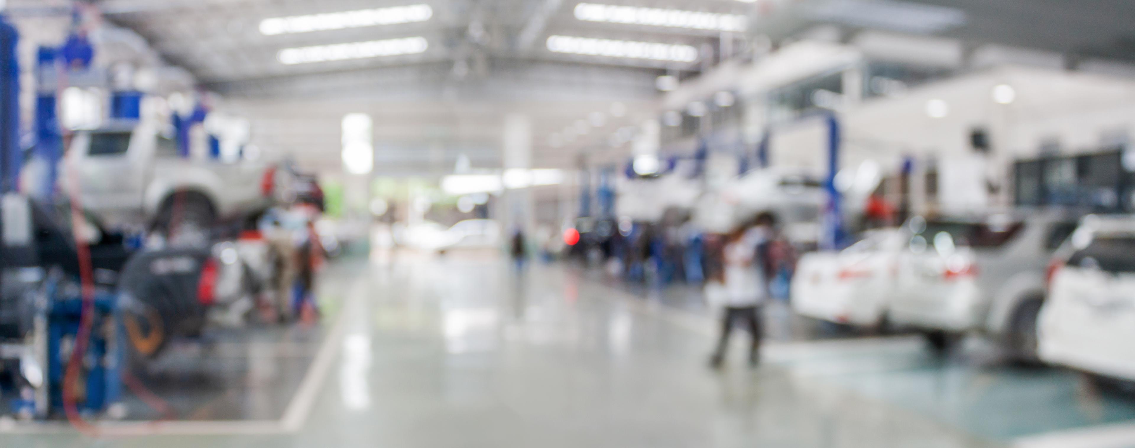 Blurred image of a large automotive workshop with cars on lifts and technicians working.