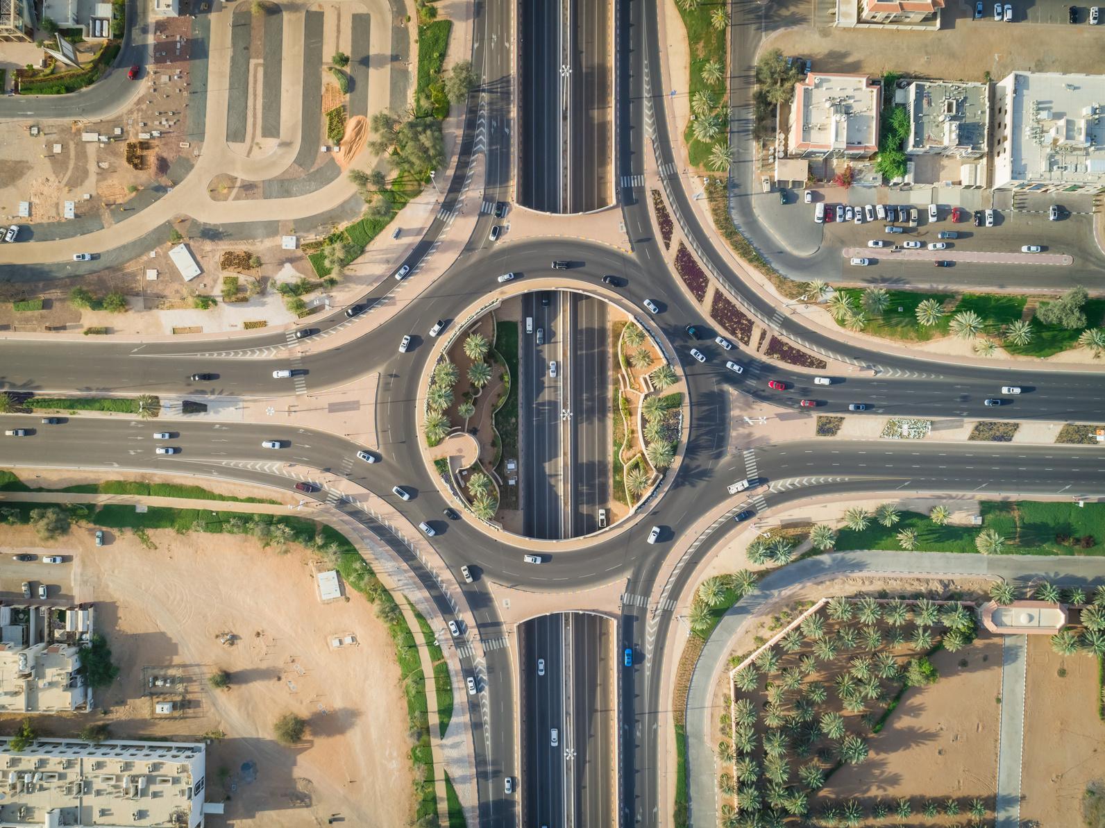 Aerial view of a large roundabout with multiple roads and vehicles, surrounded by buildings, greenery, and sandy areas.
