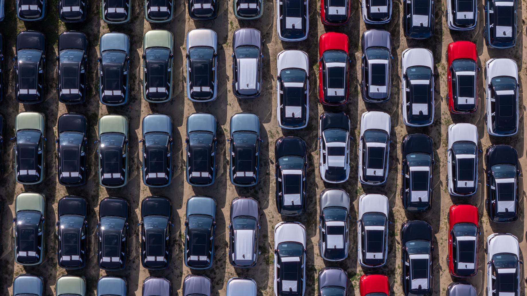 aerial-view-lined-up-cars Aerial view of a parking lot with neatly arranged rows of parked cars in various colors, including black, white, red, and blue.