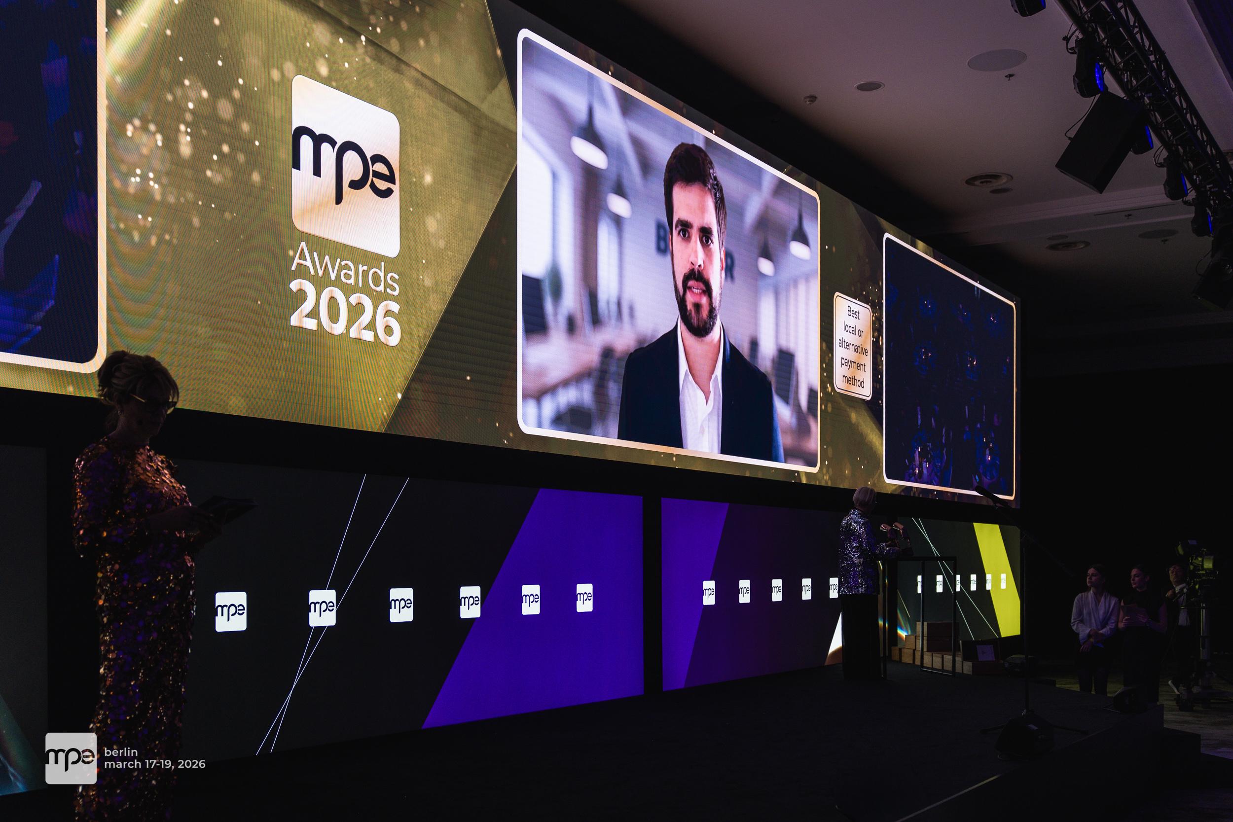 MPE_awards_2026_Best_Alternative_payment_method_winner_Bmper The MPE Awards 2026 ceremony in Berlin features a large screen with a presenter and a man on video call.
