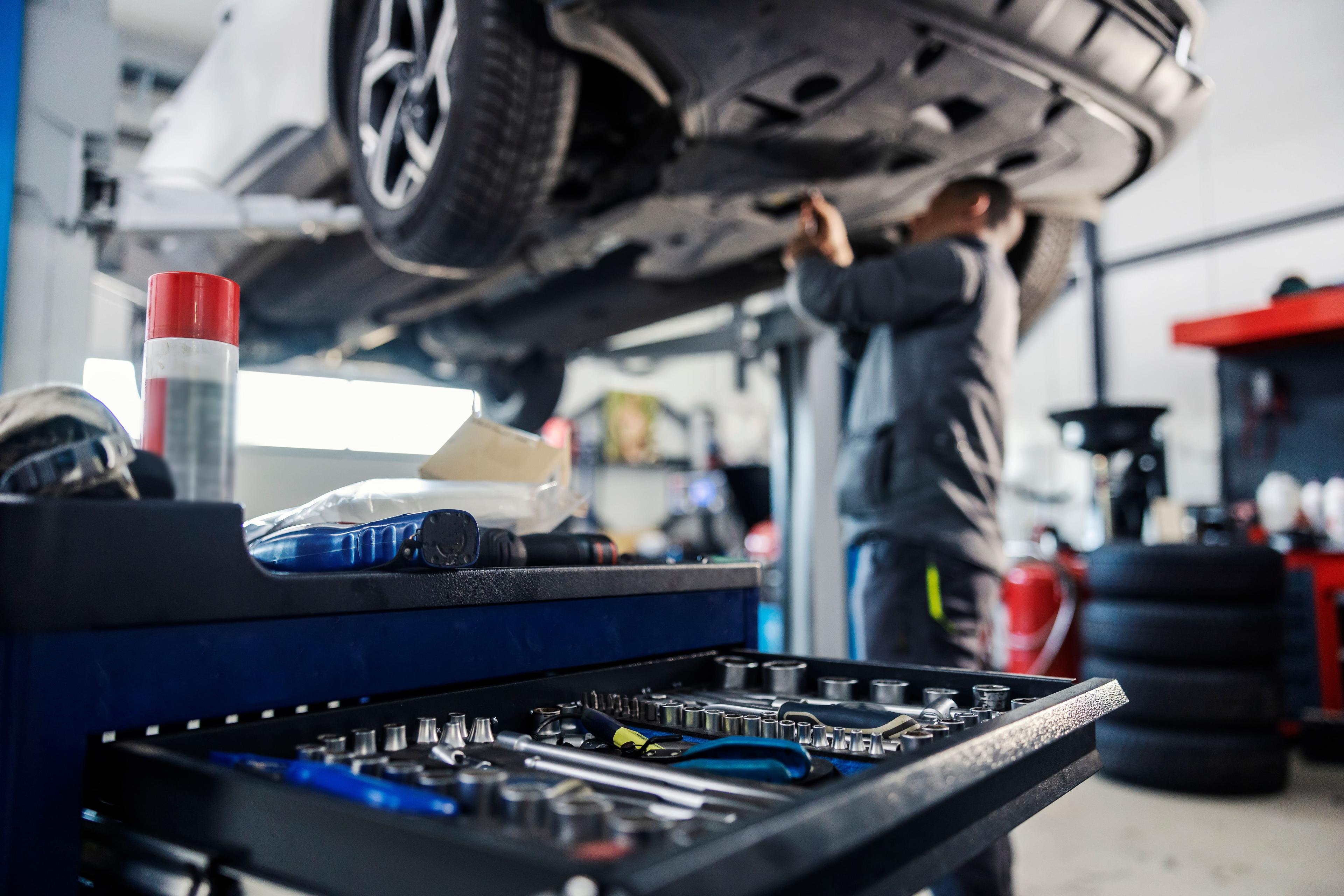 Mechanic works on a car lifted in a garage, with a focus on a tool cart filled with wrenches and other tools in the foreground.