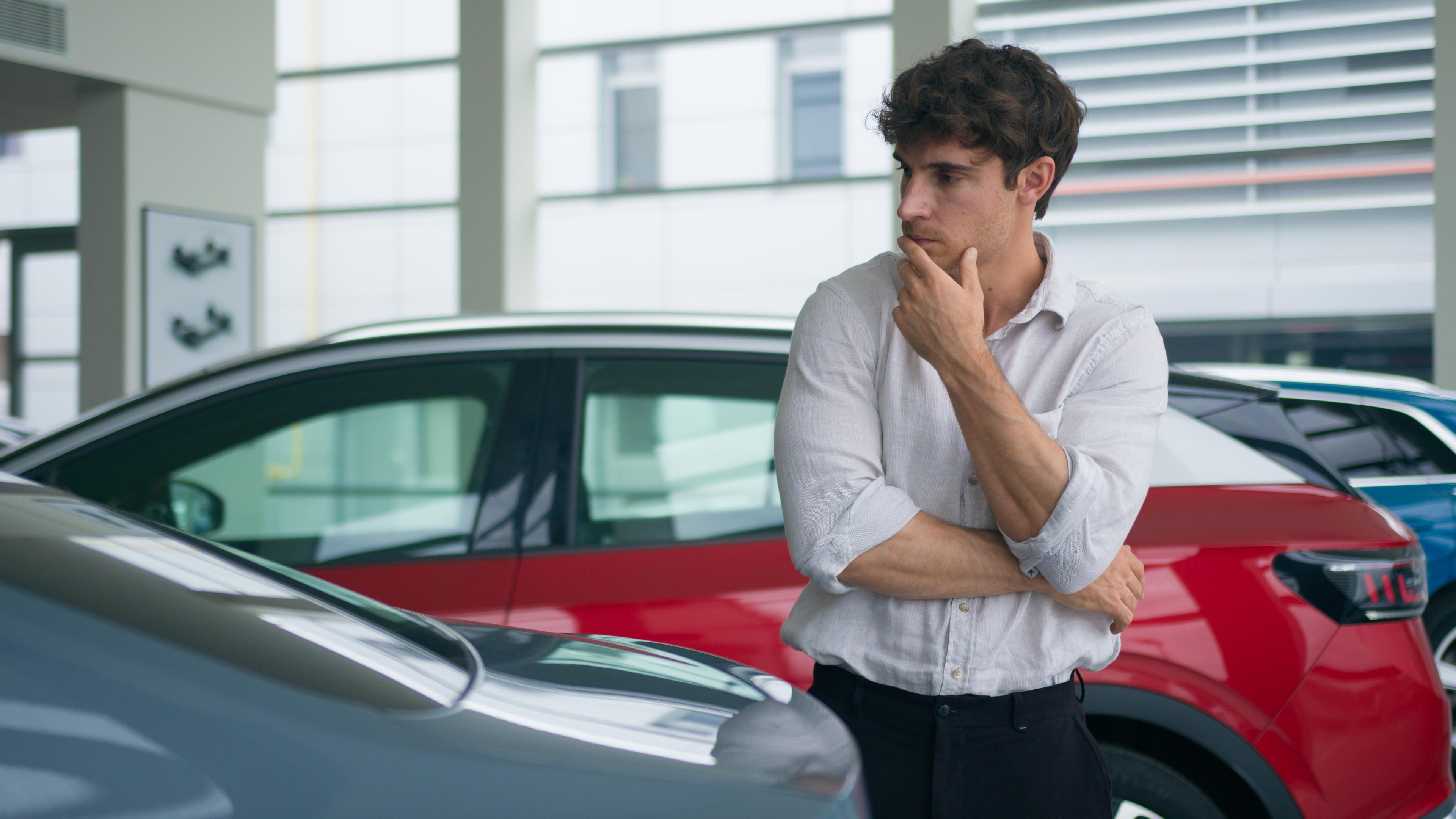 male-in-dealership-considering-wether-to-buy-or-not-a-car Man in a white shirt thoughtfully observes a car in a showroom, with a red car in the background.