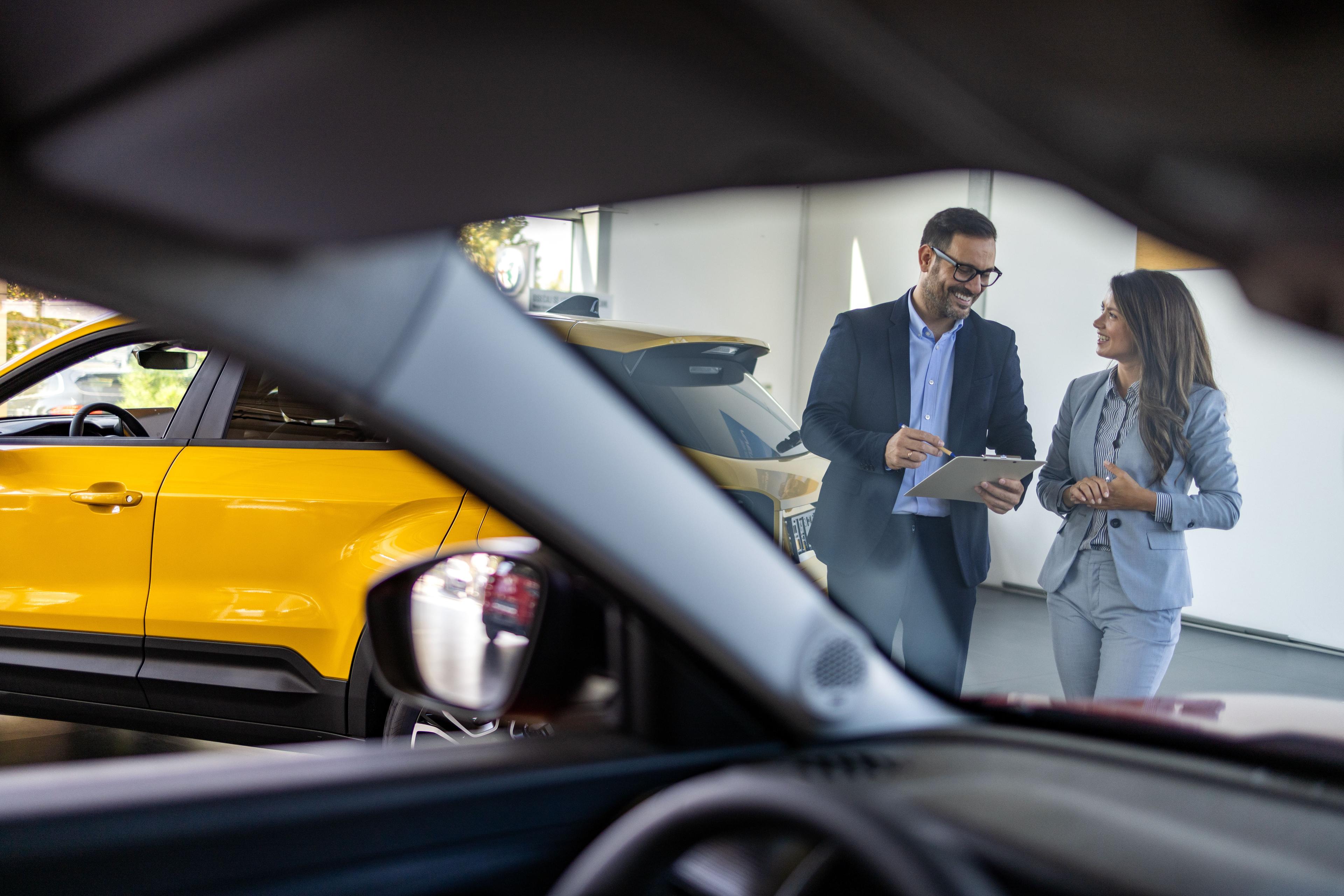 service-advisor-and-customer-conversating-seen-though-the-dashboard-of-a-car Two people in business attire stand next to a yellow car, smiling and discussing, seen through the window of another car.