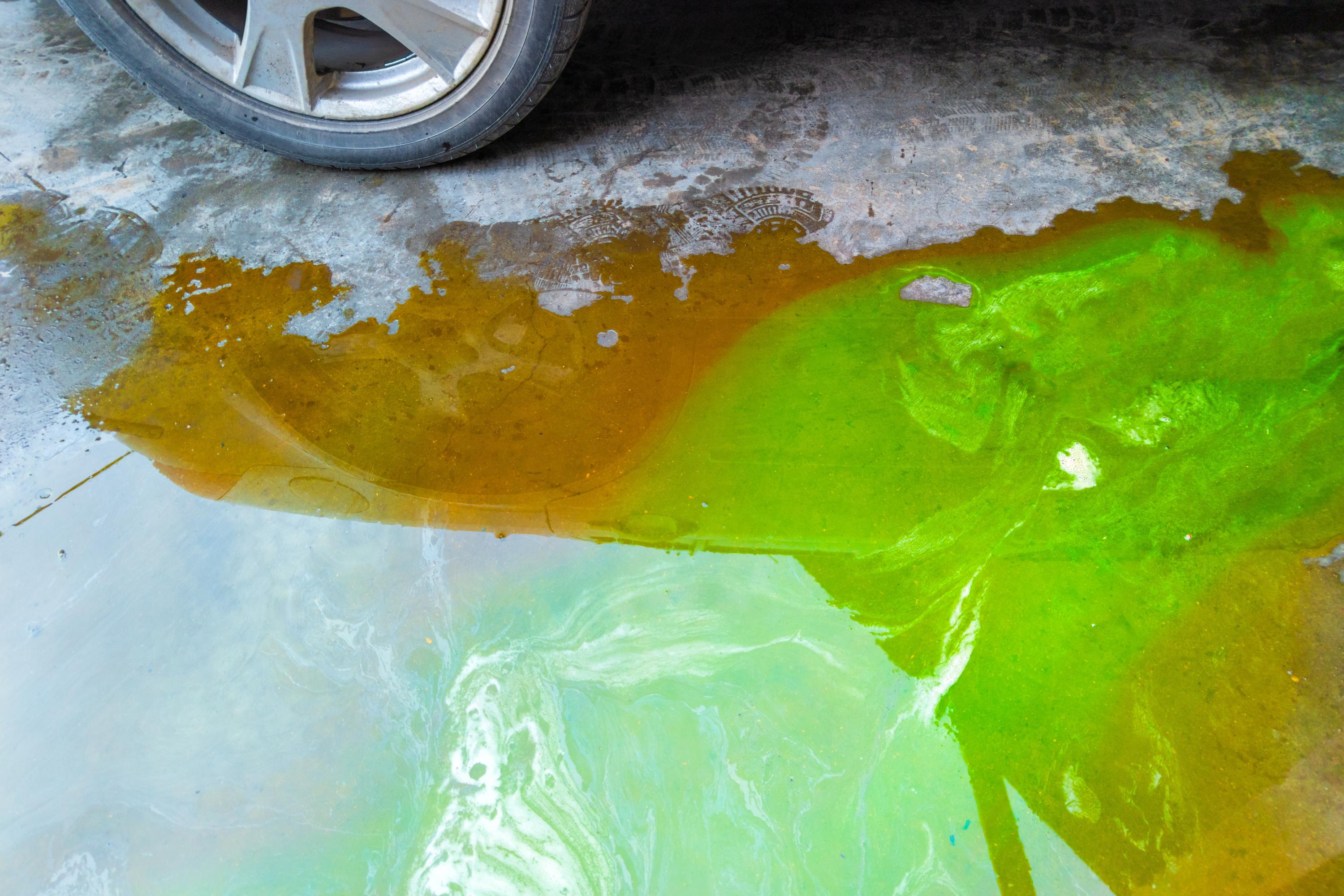 Close-up of a car tire on a wet surface with green and brown oily puddles creating a colorful pattern on the ground.